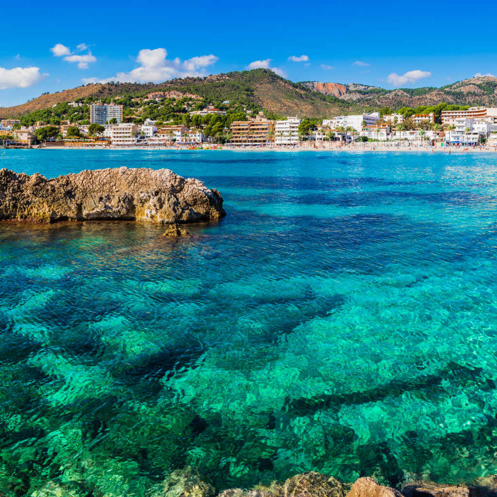 Panoramisch uitzicht op een helder turquoise zee met rotsformaties op de voorgrond en een badplaats met strandboulevard en hotelgebouwen op de achtergrond, een van de warmste winterzonbestemmingen